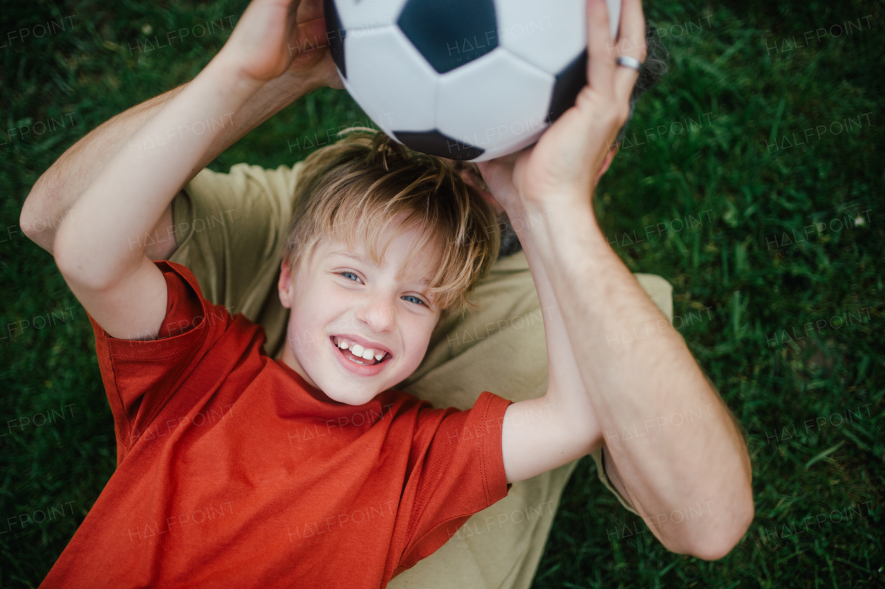 Dad lying on grass with son, laughing and having fun. Father and boy playing football, catching each other, falling to the ground. Fatherhood and Father's Day concept.