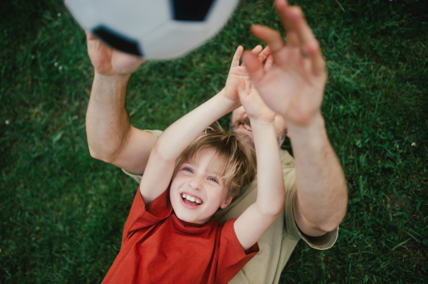 Dad lying on grass with son, laughing and having fun. Father and boy playing football, catching each other, falling to the ground. Fatherhood and Father's Day concept.