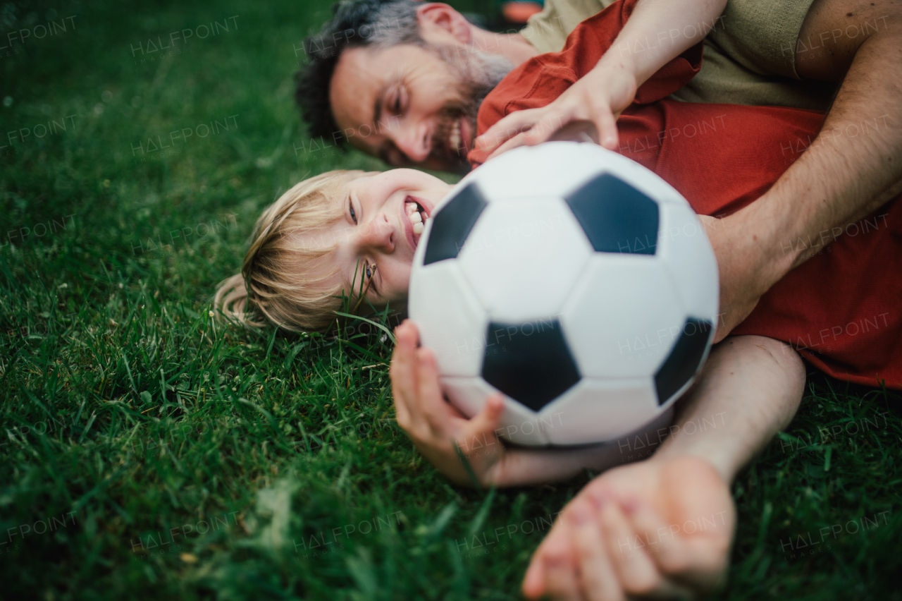 Dad lying on grass with son, laughing and having fun. Father and boy playing football, catching each other, falling to the ground. Fatherhood and Father's Day concept.