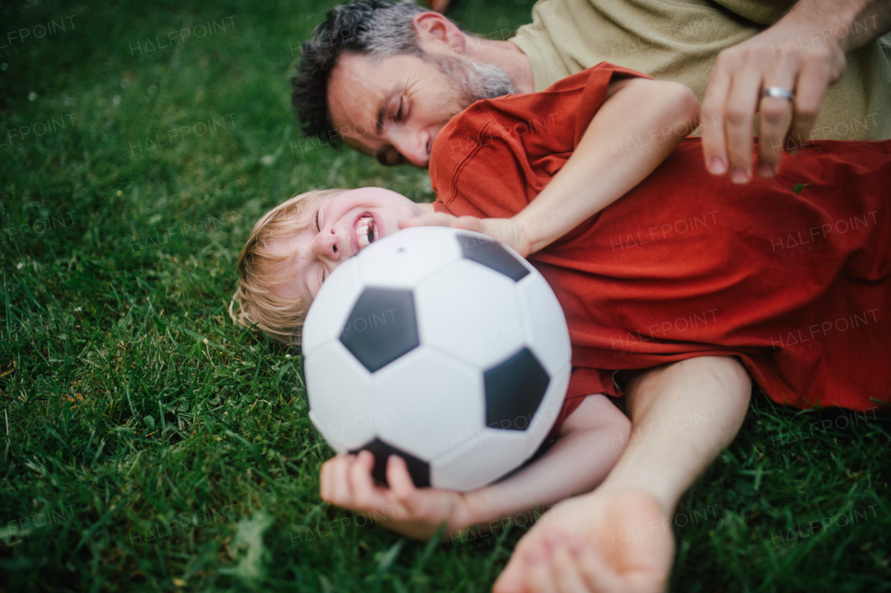 Dad lying on grass with son, laughing and having fun. Father and boy playing football, catching each other, falling to the ground. Fatherhood and Father's Day concept.