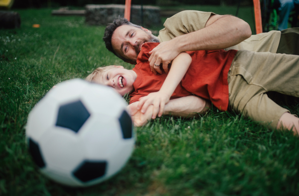 Dad lying on grass with son, laughing and having fun. Father and boy playing football, catching each other, falling to the ground. Fatherhood and Father's Day concept.