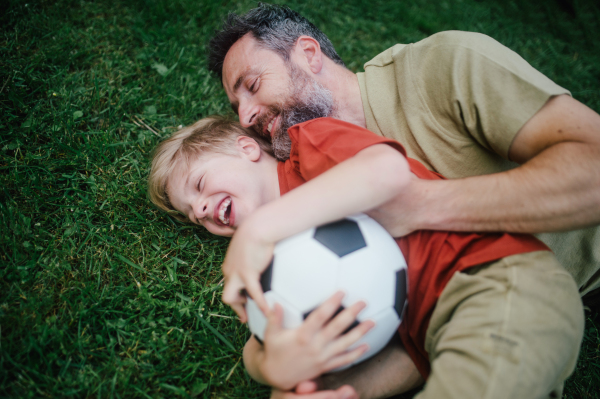 Dad lying on grass with son, laughing and having fun. Father and boy playing football, catching each other, falling to the ground. Fatherhood and Father's Day concept.