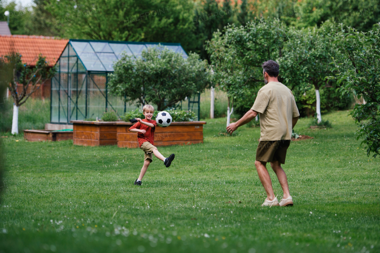 Dad having fun with young son, playing football, running and kicking ball. Playing on a lawn in front of their house. Fatherhood and Father's Day concept.