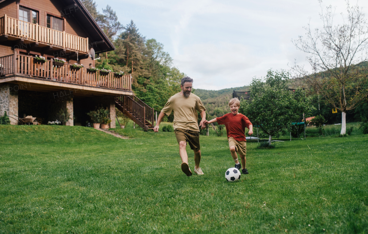 Dad having fun with young son, playing football, running and kicking ball. Playing on a lawn in front of their house. Fatherhood and Father's Day concept.