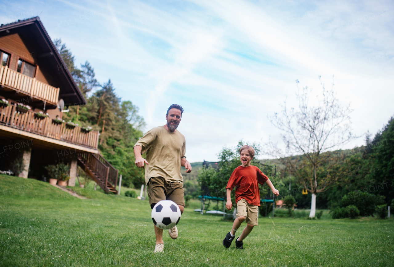 Dad having fun with young son, playing football, running and kicking ball. Playing on a lawn in front of their house. Fatherhood and Father's Day concept.