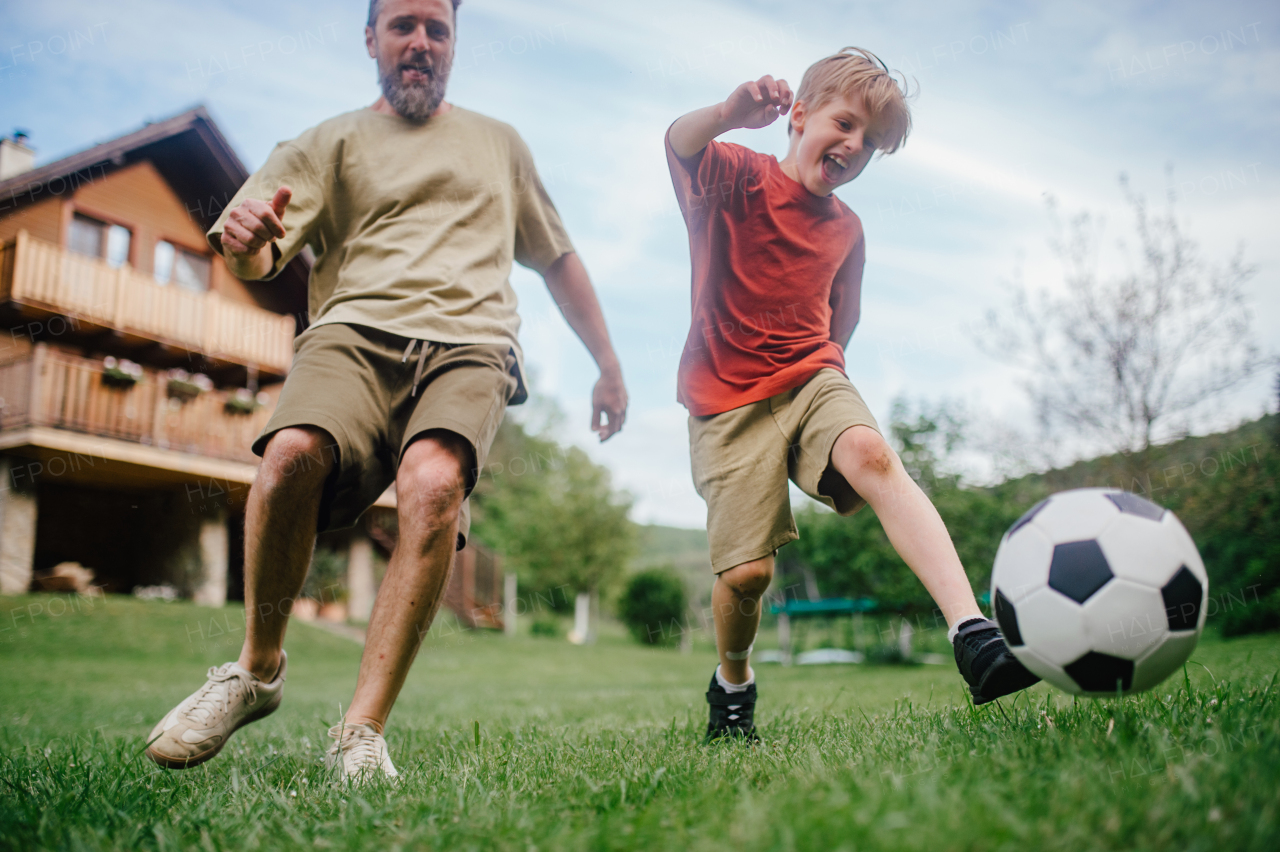 Dad having fun with young son, playing football, running and kicking ball. Playing on a lawn in front of their house. Fatherhood and Father's Day concept.