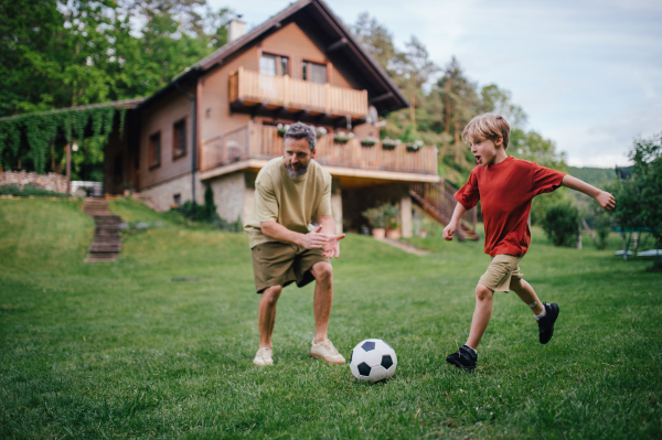 Dad having fun with young son, playing football, running and kicking ball. Playing on a lawn in front of their house. Fatherhood and Father's Day concept.