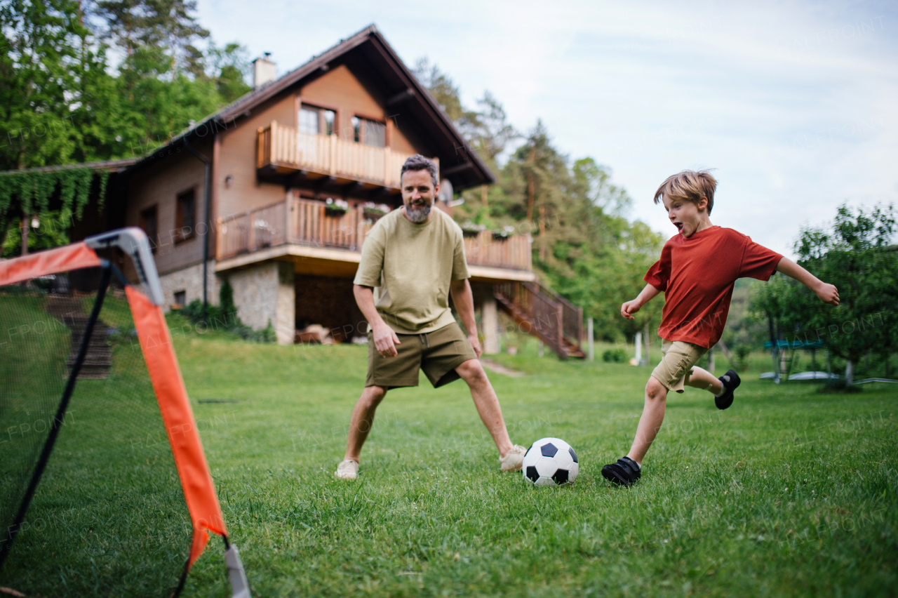 Dad having fun with young son, playing football, running and kicking ball. Playing on a lawn in front of their house. Fatherhood and Father's Day concept.