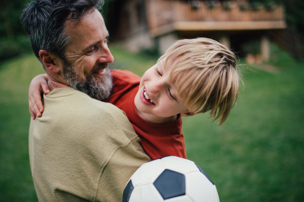 Dad embracing young son, running and celebrating the win after goal. Playing football on a lawn in front of their house. Fatherhood and Father's Day concept.