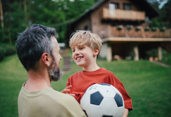 Dad embracing young son, running and celebrating the win after goal. Playing football on a lawn in front of their house. Fatherhood and Father's Day concept.