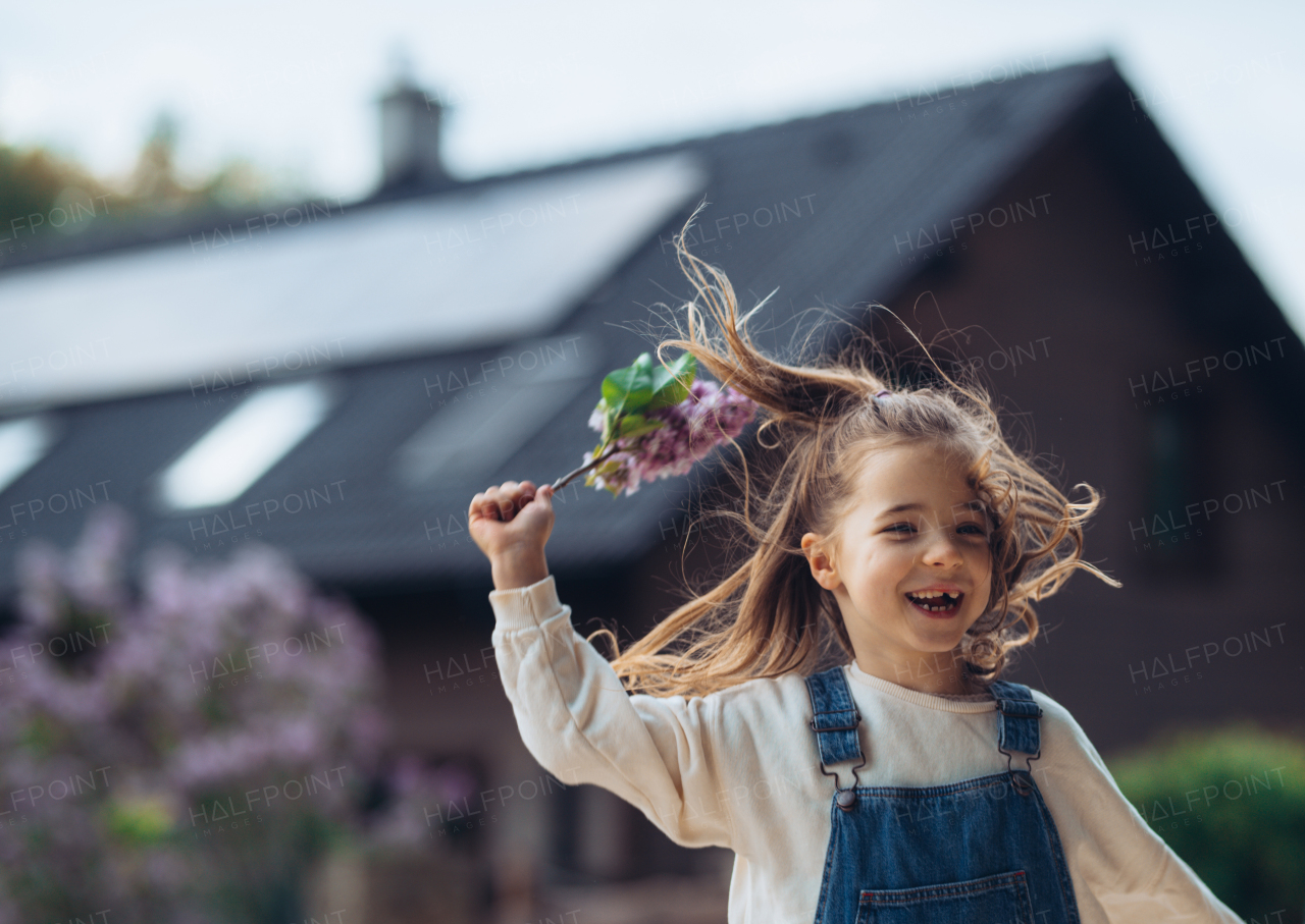 Beautiful young girl with purple lilacs flower in hands. Young girl spending time outdoors in nature, springtime, summertime. House with solar panels in background.