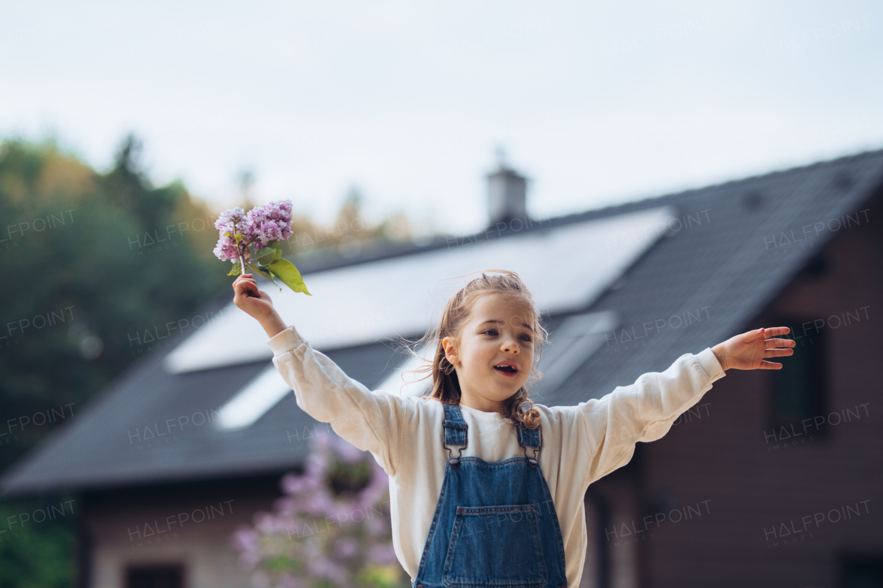 Beautiful young girl with purple lilacs flower in hands. Young girl spending time outdoors in nature, springtime, summertime. House with solar panels in background.