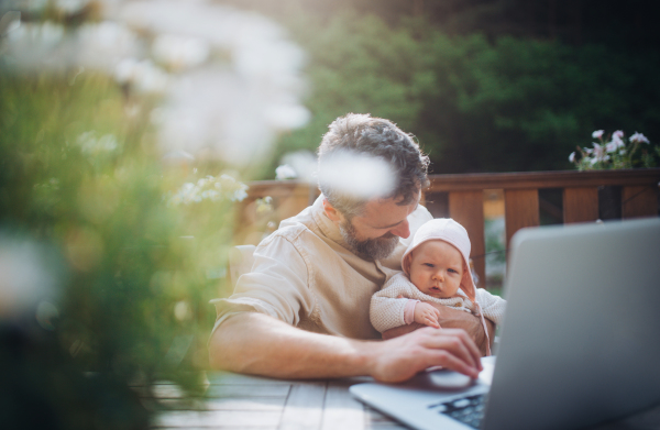 Dad working from homeoffice, sitting on patio with laptop, holding little baby girl. Father spending time with small daughter, enjoying together time. Father's Day concept.