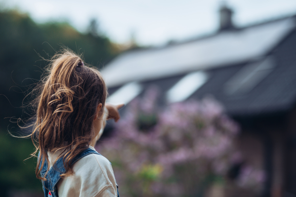 Young girl looking at house with solar panels on roof, standing outdoors in garden. Concept of springtime and solar energy.