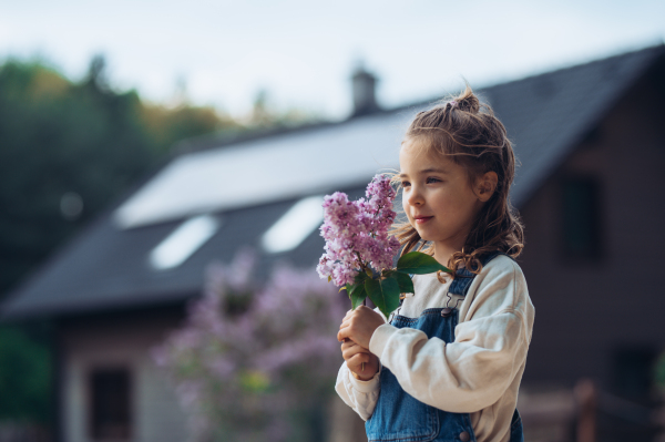 Beautiful young girl with purple lilacs flower in hands. Concept of springtime and solar energy. House with solar panels in background.