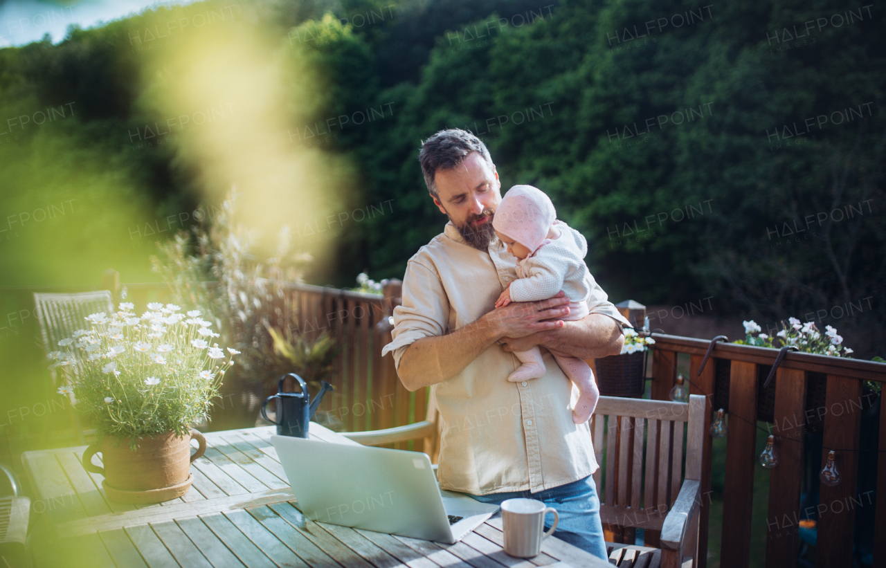 Dad taking care of his newborn baby daughter while working from homeoffice, sitting on patio with laptop.