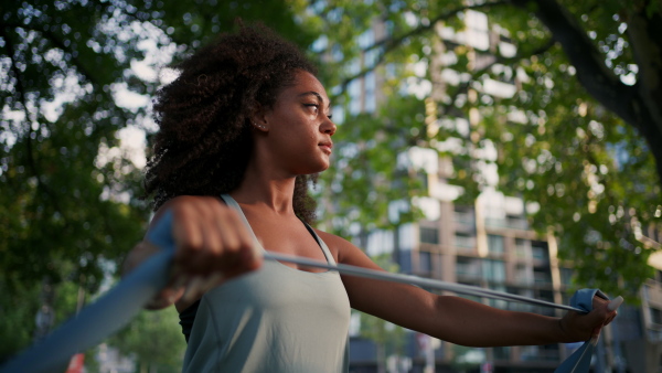 A young sporty woman exercising in city park with resistance band, stretching her arms. Doing pilates outdoors at morning.