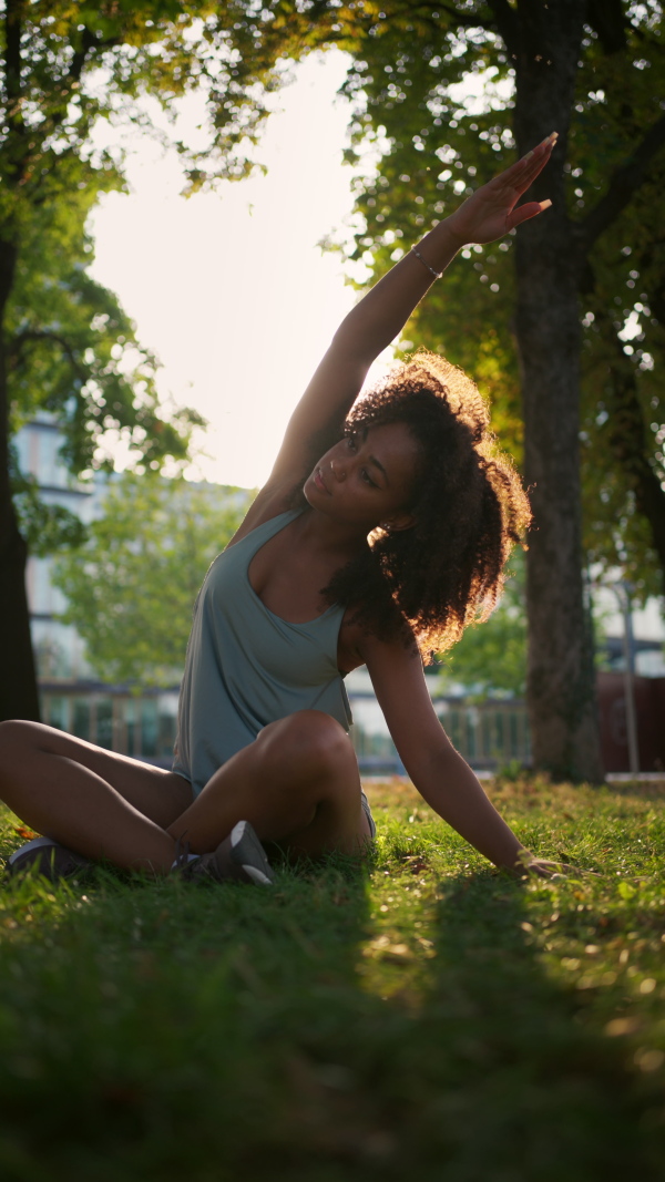 A young sporty woman exercising in city park, sitting on grass and stretching. Doing yoga outdoors at morning.