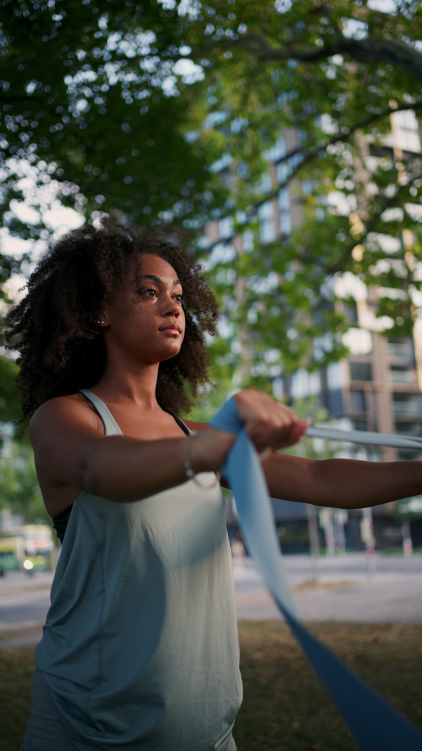 A young sporty woman exercising in city park with resistance band, stretching her arms. Doing pilates outdoors at morning.