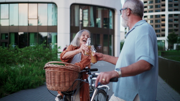 A senior couple enjoying hot summer in the city, going on bike ride around the city, drinking juice from bottle to refresh. Staycation concept.