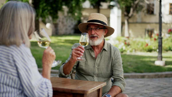 A senior couple enjoying glass of white wine, clinking with glasses. Sitting in favorite wine bar during hot summer evening in the city. Staycation concept.