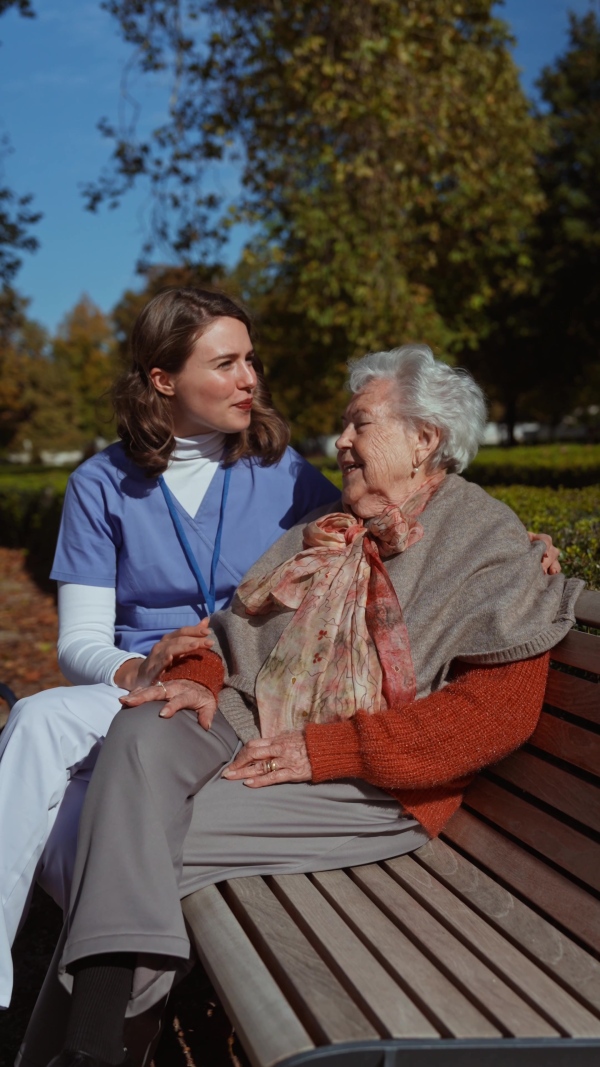 An elderly woman sitiing on bench and talking with her nurse, sharing her problems with female caregiver.