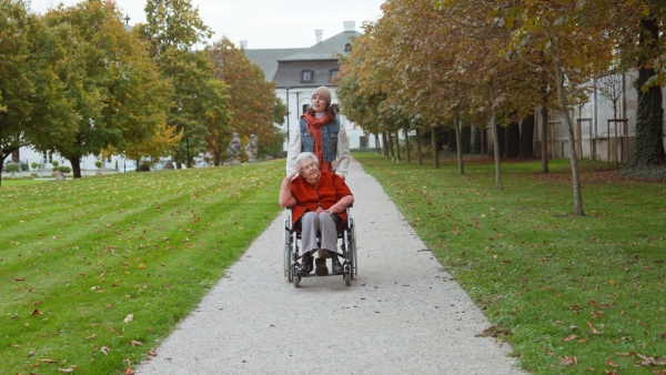 Granddaughter on an autumn walk in the park with her grandmother, pushing her in wheelchair.