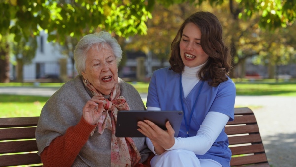 A video of nurse and elderly woman sitting on bench in park during warm autumn day. Young caregiver looking through photos of senior patient's family on tablet