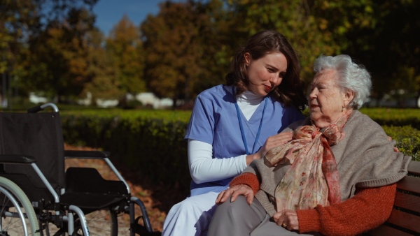 A disabled elderly woman with wheelchair sitiing on bench and talking with her nurse, sharing her problems with female caregiver.