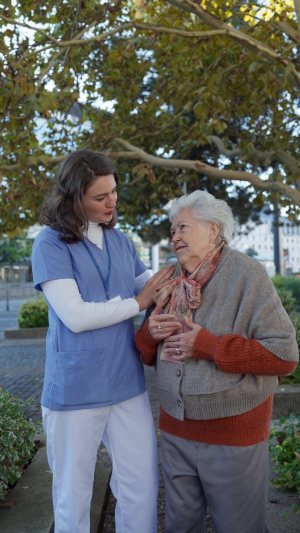 A video of nurse and elderly woman on walk in park during warm autumn day. Young caregiver spending time with senior patient.