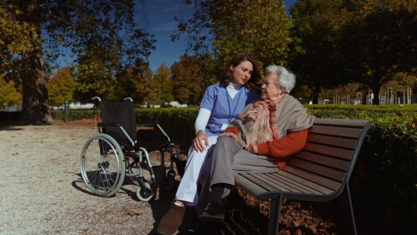 A disabled elderly woman with wheelchair sitiing on bench and talking with her nurse, sharing her problems with female caregiver.