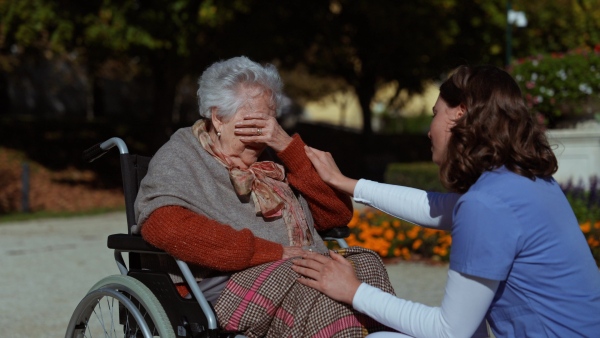 Disabled elderly woman in wheelchair talking with nurse, sharing her problems with female caregiver.