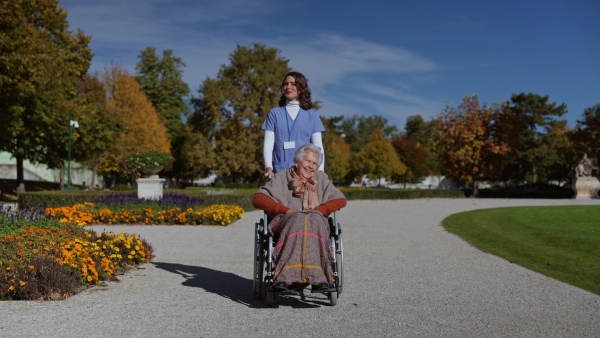 Disabled elderly woman in wheelchair on walk in park during warm autumn day. Young nurse pushing wheelchair, talking with senior woman.