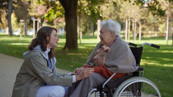 Disabled elderly woman in wheelchair talking with nurse, sharing her problems with female caregiver.