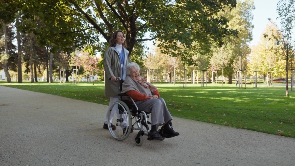 Disabled elderly woman in wheelchair on walk in park during warm autumn day. Young nurse pushing wheelchair, talking with senior woman.