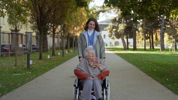 Disabled elderly woman in wheelchair on walk in park during warm autumn day. Young nurse pushing wheelchair, talking with senior woman.