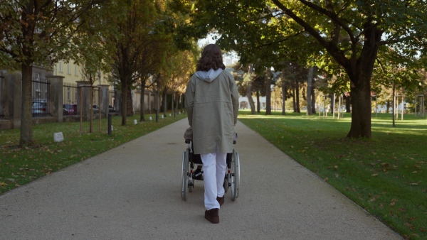 Disabled elderly woman in wheelchair on walk in park during warm autumn day. Young nurse pushing wheelchair, talking with senior woman.