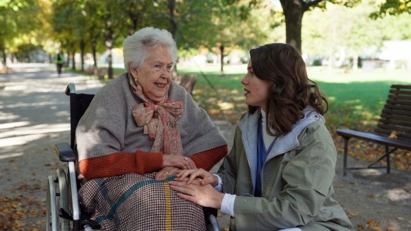 Disabled elderly woman in wheelchair talking with nurse, sharing her problems with female caregiver.