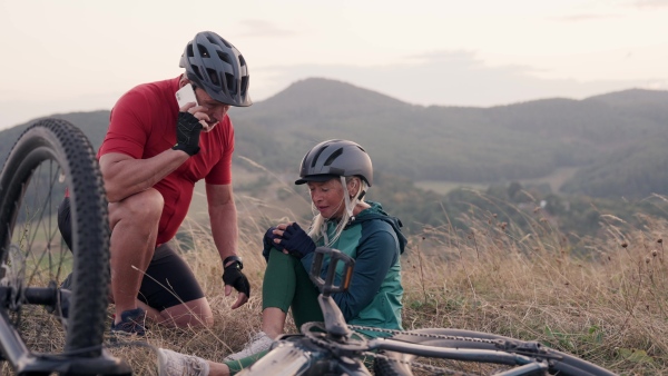 Older female cyclist fell off electric bike and injured her knee. Senior man is calling emergency with his smartphone, comforting his injured wife.