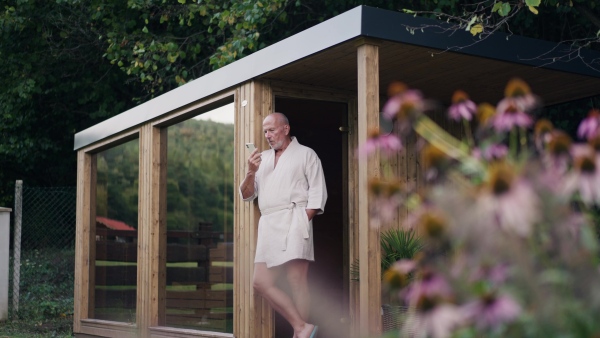 Handsome older man relaxing after spa session in home sauna, checking messages on smartphone. Concept of hygge and slow life for senior people.