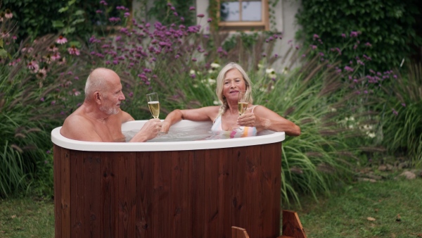 A mature couple is enjoying romantic moment in an outdoor hot tub, clinking with champagne flutes. Concept of hygge and slow life for senior people.