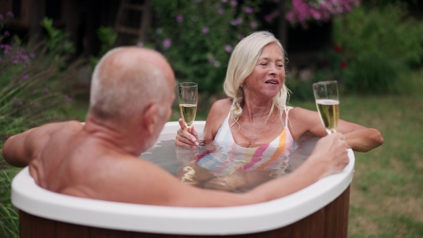 A mature couple is enjoying romantic moment in an outdoor hot tub, clinking with champagne flutes. Concept of hygge and slow life for senior people.