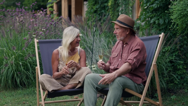 A mature couple is enjoying peaceful atmosphere in the garden, drinking champagne, sitting on folding chairs and talking. Concept of hygge and slow life for senior people.