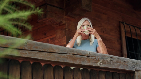 A hygge moment for beautiful senior woman standig outdoor on patio and drinking warm herbal tea.