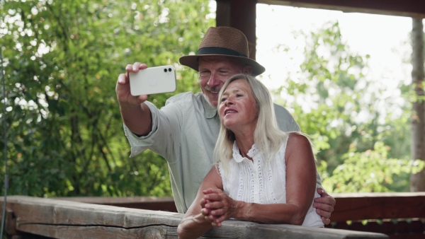 A hygge moment for beautiful senior couple standing outdoor on patio and hugging each other and taking selfie with smartphone.