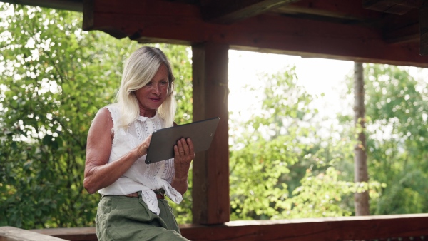 Older woman sitting outdoor on balcony or wooden patio, shopping online on tablet.