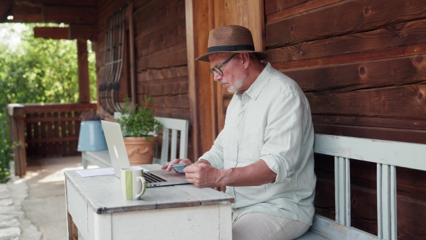 A senior man sitting outdoor on terrace, shopping online on notebook. Man holding debit card in hand, imputing card detail for payment.