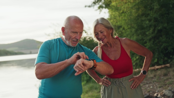 A senior couple spending morning outdoors, exercising. Senior man and woman running by lake, checking their performance on smartwatch.