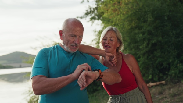 A senior couple spending morning outdoors, exercising. Senior man and woman running by lake, checking their performance on smartwatch.