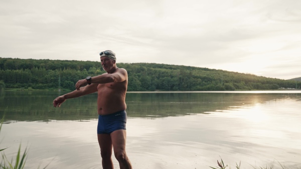 A senior man has a shared hobby, swimming in lake during cold autumn morning. Stretching before going into water.